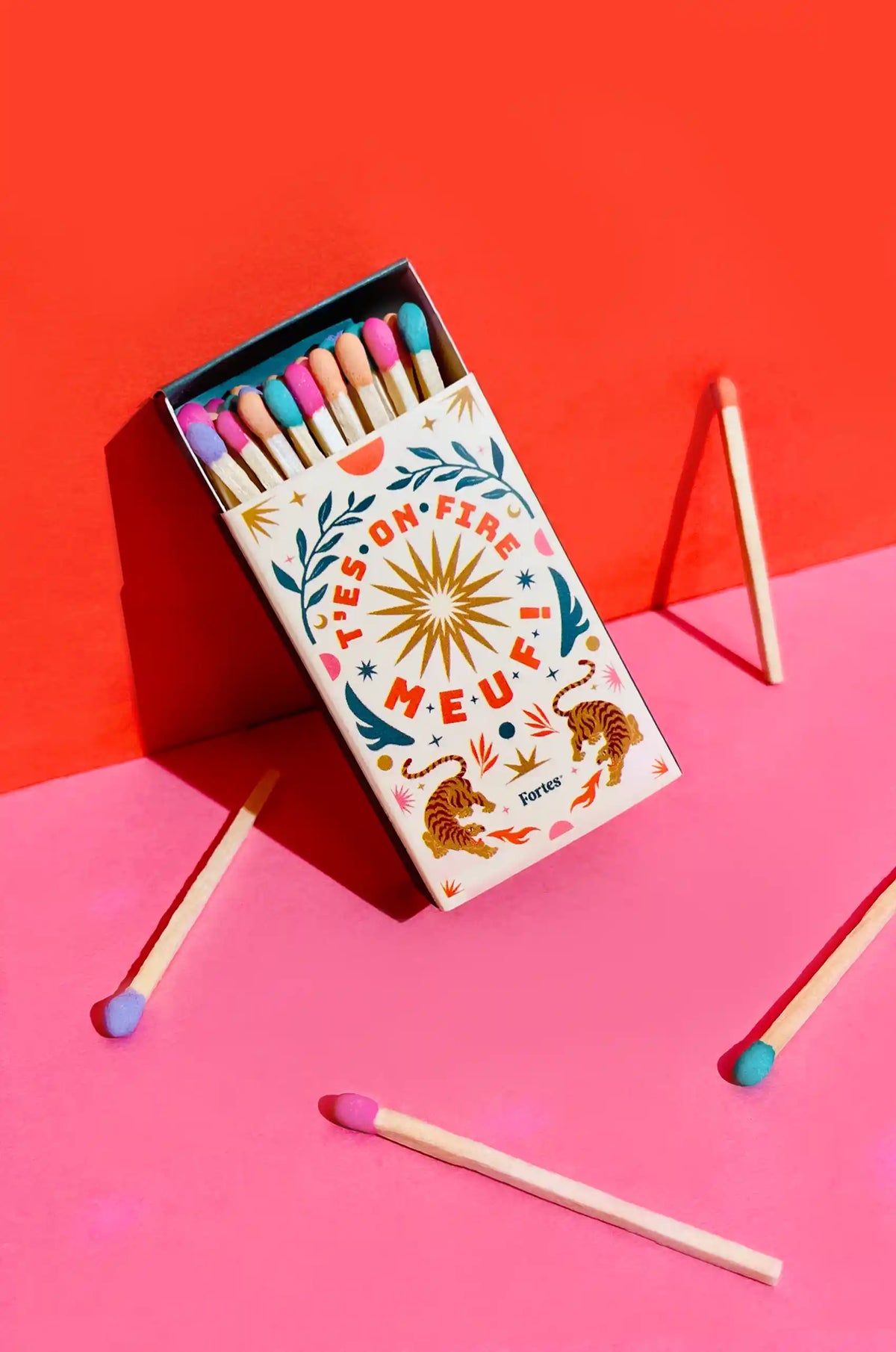 Petite boîte d’allumettes multicolores Fortes ouverte sur fond rouge, avec inscription « T’es on fire meuf »