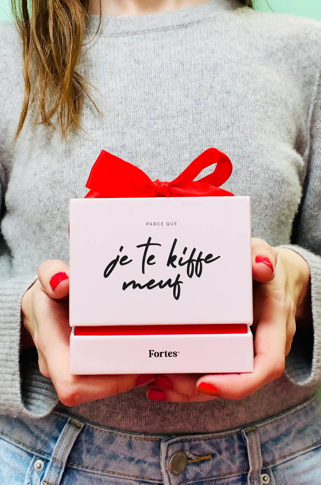 Femme tenant un coffret rose Fortes « Je te kiffe meuf », fermé par un nœud rouge